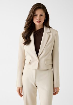 Frau mit langem braunem Haar trägt einen beigen, maßgeschneiderten Blazer und passende, hochtaillierte Hosen über einem dunkelbraunen Oberteil und steht vor einem weißen Hintergrund.