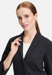 Zwarte gestructureerde blazer met een geribbelde kraag, twee knopen en een textuurstof. Draag het over een witte top, wat een getailleerde stijl laat zien.