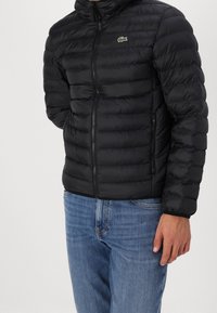 Černá péřová bunda s kapucí, prošitým designem, zapínáním na zip a zeleným logem na hrudi. Nasažená přes modré džíny.