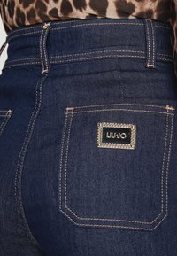 Jeans de mezclilla oscura con cintura alta, costuras doradas y un bolsillo trasero con un detalle metálico rectangular del logo de LIU JO.