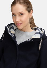 Ung kvinde med lyst hår iført en navy frakke med hætte med mønster og en lys grå sweater, der smiler blidt mod en hvid baggrund.