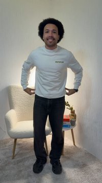 Άνδρας με αφροχτένισμα, φορώντας λευκό φούτερ Napapijri και μαύρο παντελόνι, στέκεται και χαμογελά σε ένα ζεστό δωμάτιο με μια καρέκλα και ένα μικρό τραπέζι.