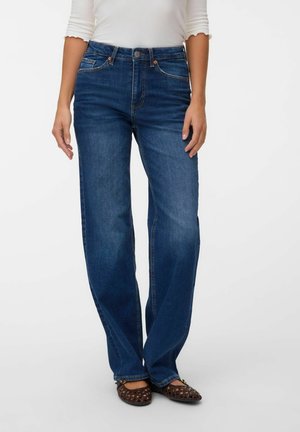 Personne portant un jean droit bleu foncé, un haut blanc à manches trois-quarts et des chaussures plates tressées marron sur fond blanc.