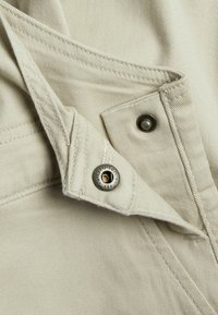 Close-up van beige stof met gedetailleerd stiksel en een gedeeltelijk geopende metalen drukknoop op de klep van een kledingzak.