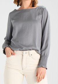 Blusa grigia a maniche lunghe in tessuto leggero, con scollatura a barca e polsini arricciati. Abbinata a pantaloni chiari a vita alta.