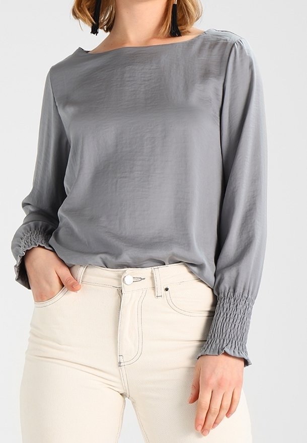 Blusa grigia a maniche lunghe in tessuto leggero, con scollatura a barca e polsini arricciati. Abbinata a pantaloni chiari a vita alta.