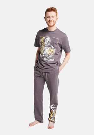 Junger Mann steht barfuß, trägt ein graues Mandalorianer-Themen-T-Shirt und passende Hose, hat die Hände in den Taschen und lächelt in die Kamera.