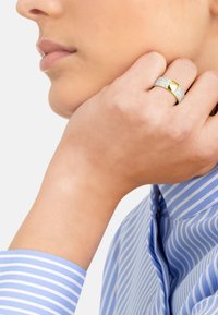 Anello d'oro con una banda testurizzata caratterizzata da accenti scintillanti e un dettaglio centrale liscio. Indossato su una mano vicino a una camicia a righe blu.