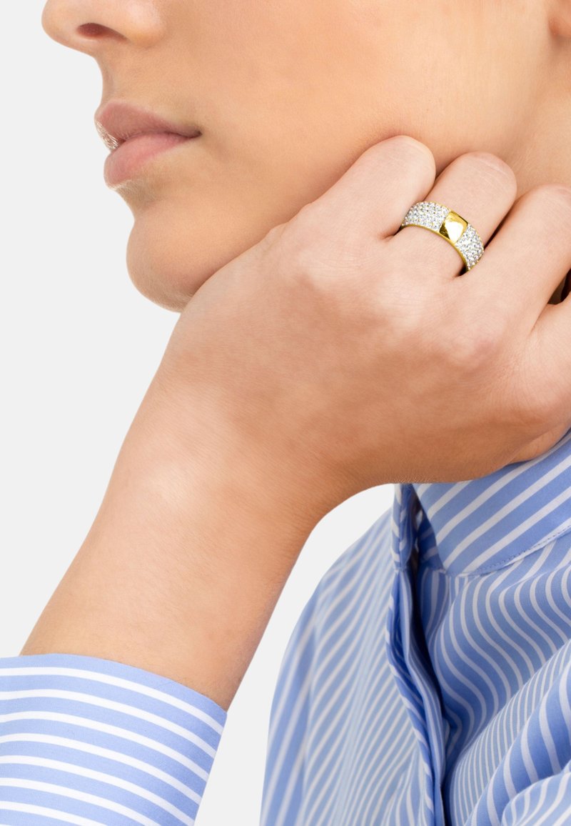 Anello d'oro con una banda testurizzata caratterizzata da accenti scintillanti e un dettaglio centrale liscio. Indossato su una mano vicino a una camicia a righe blu.