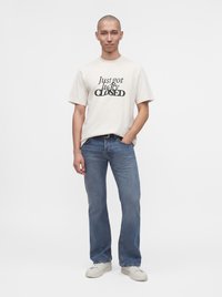 Homme debout avec une main dans la poche, portant un t-shirt blanc avec un texte et un jean bleu sur un fond uni.