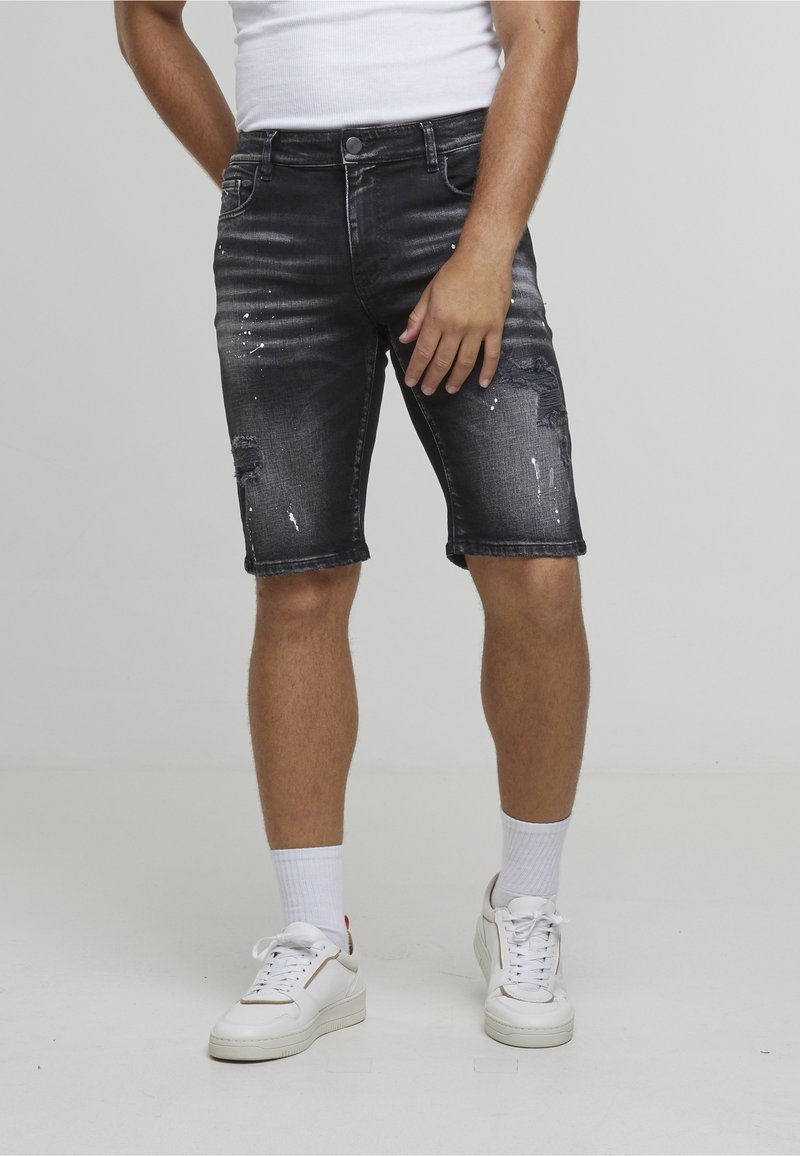 Homme portant un short en denim noir usé, des chaussettes blanches, des baskets blanches et un haut blanc sans manches, debout devant un fond uni.