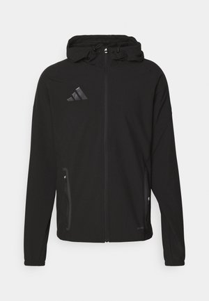Schwarze sportliche Jacke mit Kapuze, ausgestattet mit einem Reißverschluss vorne, zwei seitlichen Taschen und einem glänzenden Logo auf der linken Brust. Glatte Stoffoberfläche.