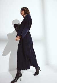 Robe longue marine en tissu texturé, avec des manches larges et un ourlet évasé, associée à des bottines noires et un sac noir, sur fond blanc.