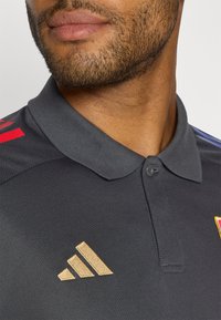 Donkergrijze polo met een textuurstof, voorzien van rode strepen en een gouden Adidas-logo op de linkerkant van de borst.