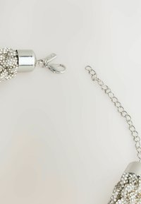 Sudraba kulons ar teksturētu virsmu, izrotāts ar mazām caurspīdīgām akmeņiem, ar metāla augšdaļu un ķēdi, kas piestiprināta ar aizdari.