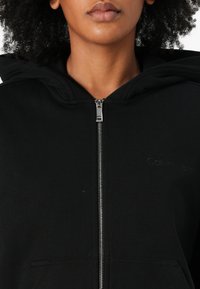 Čierna mikina na zips vyrobená z mäkkej látky, ktorá má predný zips, dve vrecká a diskrétne logo na oblasti hrude. S kapucňou.