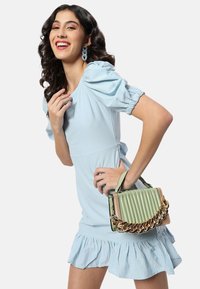 Ljusblå klänning med puffärmar och volangkant; håller en grön, ribbad handväska med guldkedjeaccenter. Slät tygkvalitet.