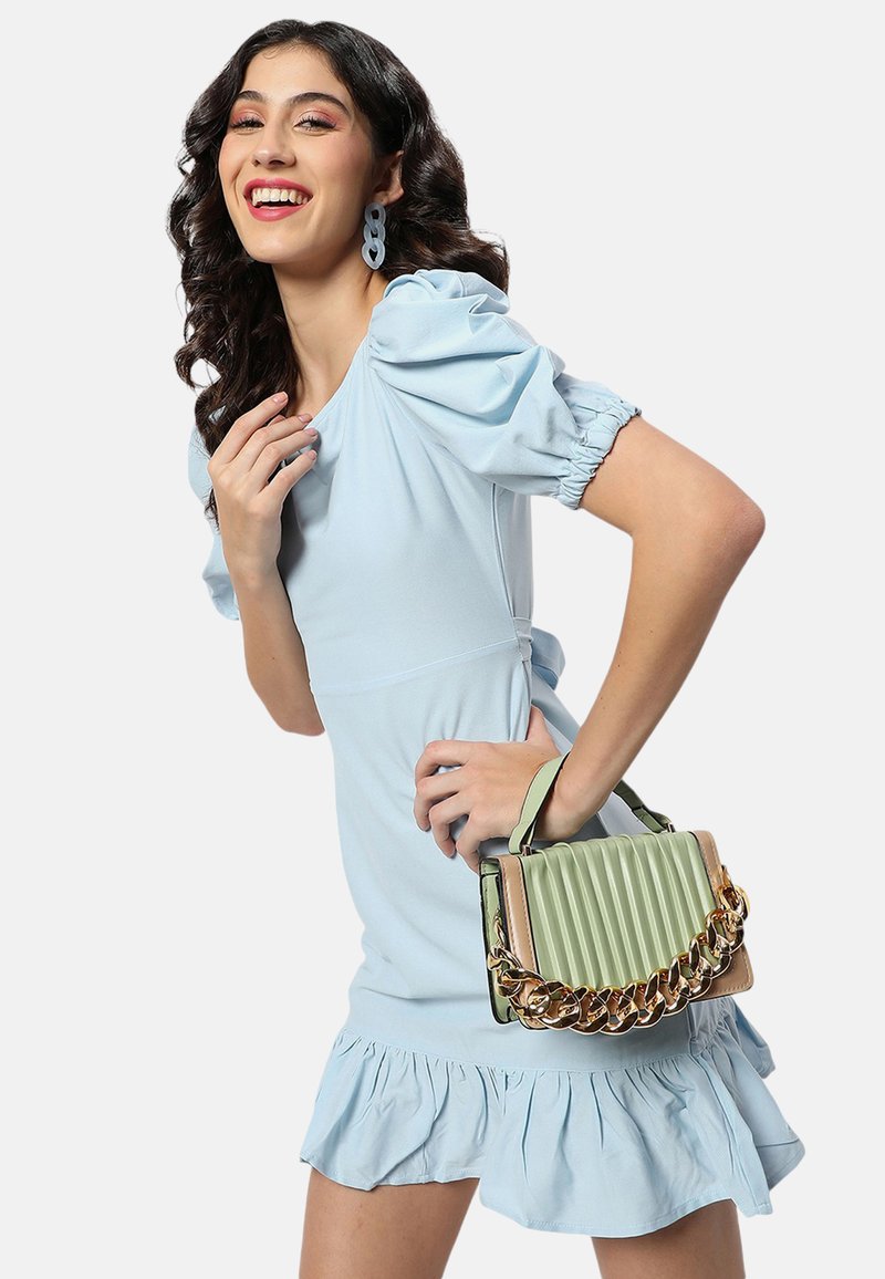 Ljusblå klänning med puffärmar och volangkant; håller en grön, ribbad handväska med guldkedjeaccenter. Slät tygkvalitet.