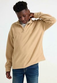 Beige fleece pullover with a half-zip neck, long sleeves, and a relaxed fit, paired with blue jeans, creating a casual look.