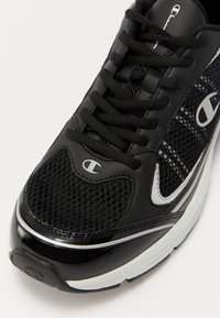 Black athletic sneaker featuring a mesh textile upper, synthetic leather accents, round laces, and a curved white sole with branding.