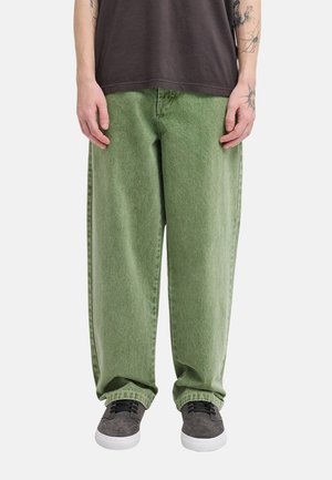 Osoba ubrana w luźne zielone dżinsy, szare trampki i ciemny t-shirt z widocznymi tatuażami na przedramionach na jednolitym tle.