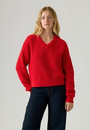 Czerwony sweter z żebrowanym splotem, z dekoltem w kształcie litery V i obniżonymi ramionami, połączony z granatowymi dżinsami o wysokim stanie. Miękka tekstura i luźny krój.