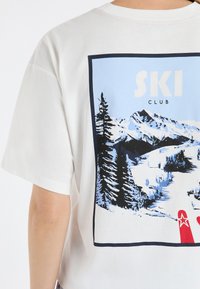 Biała bawełniana koszulka z niebieskim nadrukiem graficznym przedstawiającym góry i drzewa oraz napisem "SKI CLUB". Krótkie rękawy i luźny krój.