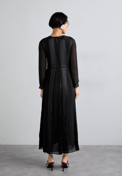 Robe noire à manches longues avec un overlay en tissu transparent, design plissé, taille cintrée et détail en forme de clef à l'arrière. Chaussures à petits talons visibles.