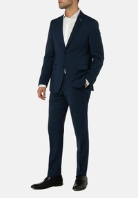 Man in een nauwsluitend marineblauw pak met een wit overhemd en zwarte nette schoenen, staande met één hand in zijn zak.