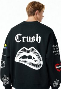 Mladý muž stojaci chrbtom, oblečený v čiernej mikine s veľkými bielymi pierkami a slovom "Crush", doplnený rôznymi farebnými symbolmi a textom na rukávoch.