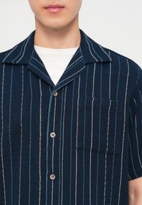 Άνδρας που φοράει μπλε σκούρο κοντομάνικο πουκάμισο με κάθετες λευκές και ανοιχτό μπλε ρίγες πάνω από λευκό μπλουζάκι.