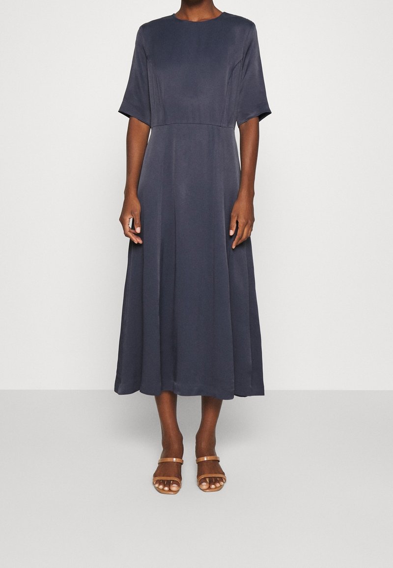 Femme portant une robe midi bleu marine foncé à manches courtes et taille cintrée, associée à des sandales à bout ouvert couleur beige.