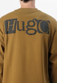 Brown sweatshirt featuring a large gray "Hugo" logo on the back, made from soft fabric with a smooth texture and a relaxed fit.