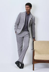 Helles graues Anzugensemble mit einem taillierten Blazer und einer Hose, kombiniert mit einem dunkel gemusterten Hemd. Auffällige Details sind ein Einstecktuch und elegante schwarze Schuhe.