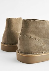 Botas de tobillo de ante marrón con forma redondeada, que presentan costuras visibles y una superficie texturizada, montadas sobre una suela de goma plana.