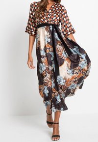 Donna che indossa un vestito avvolgente con motivo, con parte superiore geometrica e gonna floreale, cintura nera in vita e tacchi aperti neri con cinturino alla caviglia mentre cammina.