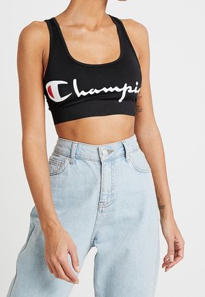 Damski tors w czarnym sportowym biustonoszu Champion i jasnoniebieskich jeansach z wysokim stanem, z widocznymi tatuażami na ramionach.