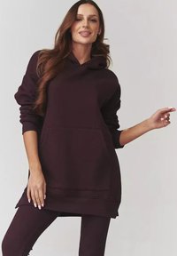Bordowy hoodie z kapturem, z prążkowanymi mankietami, przednią kieszenią i dłuższym dołem. Noszony z dopasowanymi prążkowanymi legginsami. Gładka tekstura.