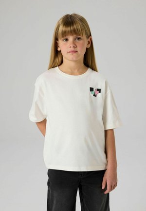 Camiseta blanca de algodón con mangas cortas, que presenta un pequeño gráfico de corazón en negro, verde y rosa en el área del pecho izquierdo. Combinada con pantalones negros.