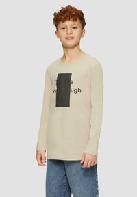 Beige longsleeveshirt met een ronde halslijn, voorzien van een zwart rechthoekig grafisch ontwerp met tekst. Gecombineerd met lichtblauwe denim jeans.