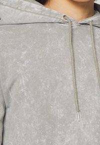 Felpa grigia con superficie testurizzata, dotata di cappuccio con cordino e accenti metallici. Design minimalista con leggere variazioni di colore.