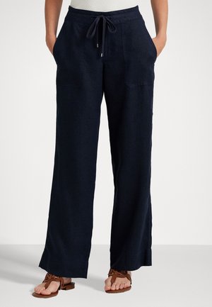 Vrouw draagt marineblauwe wijde broek met voorzakken en een taille met koordje, gecombineerd met bruine platte sandalen.