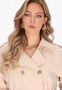 Trench coat beige con collare a revers, bottoni dorati doppi, collana a catena dorata a strati e capelli biondi mossi.