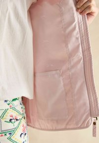 Innenfutter einer hellrosa Jacke mit einer Tasche, glatter samtartiger Textur und subtil geprägten Mustern von Pferden. Ungefütterte Kanten und Reißverschluss.