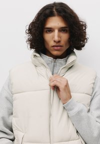 Beige puffer vest with a high collar, zip closure, and horizontal stitching, layered over a light gray sweatshirt with ribbed cuffs.