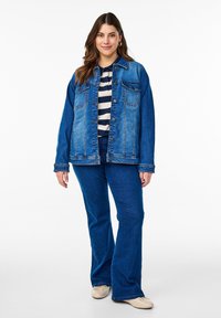 Traper jakna s opuštenim krojem, dva džepa na prsima i gumbima. Uparen s prugastom majicom i plavim trapericama širokih nogavica. Svijetla pozadina.