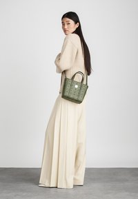 Πράσινη τσάντα υφής με κοντούς χειρολισθήρες, διακοσμημένη με στρογγυλή λογότυπο. Το μοντέλο φοράει ένα μπεζ πλεκτό πουλόβερ και κρεμ παντελόνια με φαρδιά πόδια.