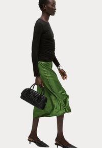 Schwarzes Langarmoberteil, grüner Satin-Midi-Rock, schwarze strukturierte Handtasche mit geprägtem Detail und schwarze Kitten-Heel-Mules.
