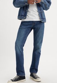 Personne portant un jean droit bleu foncé, des baskets noires et blanches, une chemise blanche et une veste en jean bleue, les mains ajustant le devant de la veste.