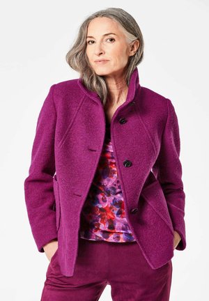 Manteau en mélange de laine violet avec un col haut et des boutons noirs, porté sur un haut à motifs colorés, associé avec un pantalon violet assorti.
